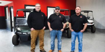 Three middle-aged men stand in a row facing the camera with their hands in their pockets inside a vehicle showroom. The man on the left wears glasses, a black hoodie, tan pants, and work boots. The center man wears a black baseball cap, a black polo shirt, blue jeans, and brown boots. The man on the right wears a black polo shirt, blue jeans, and gray slip-on shoes. Both polo shirts and the hoodie feature a small red and white "GT" logo on the left chest. Behind them, three modern golf carts—one dark green, one tan, and one white—are parked on a gray speckled epoxy floor against a vibrant red and dark gray wall.