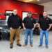 Three middle-aged men stand in a row facing the camera with their hands in their pockets inside a vehicle showroom. The man on the left wears glasses, a black hoodie, tan pants, and work boots. The center man wears a black baseball cap, a black polo shirt, blue jeans, and brown boots. The man on the right wears a black polo shirt, blue jeans, and gray slip-on shoes. Both polo shirts and the hoodie feature a small red and white "GT" logo on the left chest. Behind them, three modern golf carts—one dark green, one tan, and one white—are parked on a gray speckled epoxy floor against a vibrant red and dark gray wall.