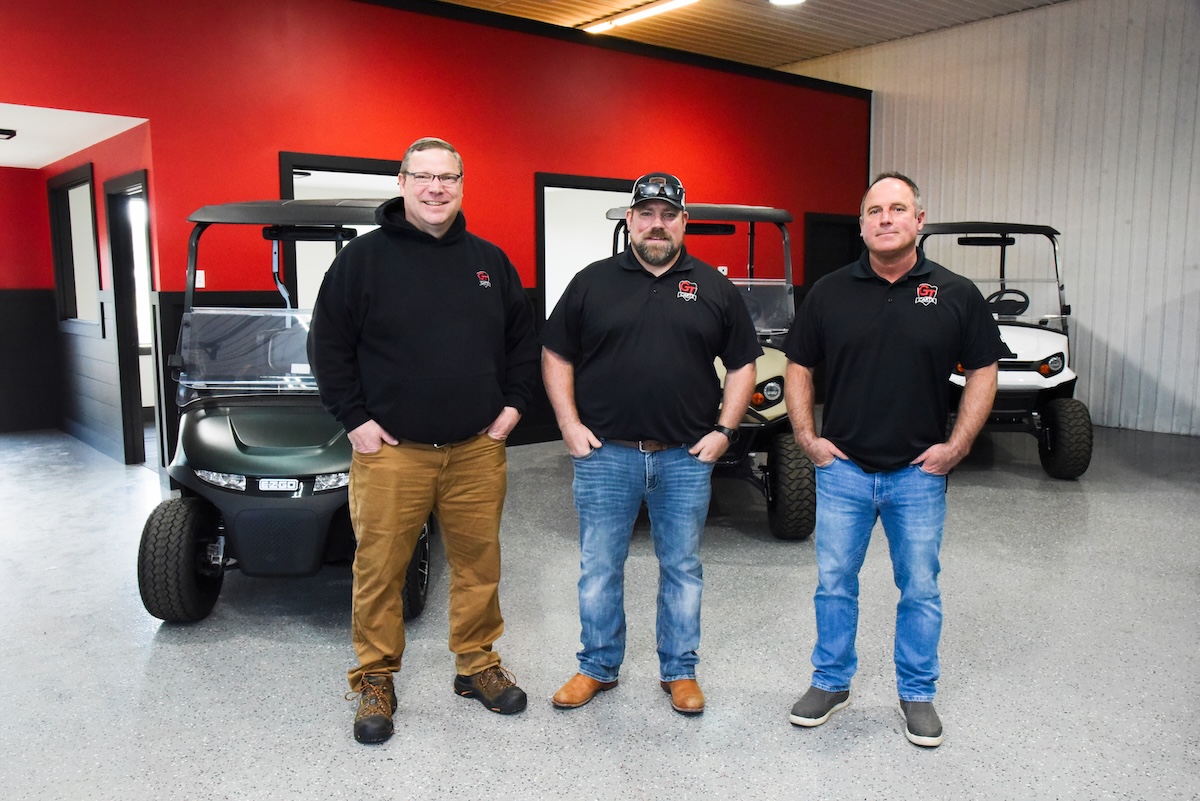 Three middle-aged men stand in a row facing the camera with their hands in their pockets inside a vehicle showroom. The man on the left wears glasses, a black hoodie, tan pants, and work boots. The center man wears a black baseball cap, a black polo shirt, blue jeans, and brown boots. The man on the right wears a black polo shirt, blue jeans, and gray slip-on shoes. Both polo shirts and the hoodie feature a small red and white "GT" logo on the left chest. Behind them, three modern golf carts—one dark green, one tan, and one white—are parked on a gray speckled epoxy floor against a vibrant red and dark gray wall.
