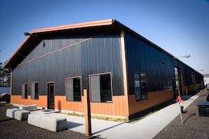 A wide-angle exterior view of a large, contemporary metal facility on a sunny day. The building features vertical corrugated metal siding, with the top portion painted dark gray and the bottom portion painted a warm copper-orange color. The front of the structure has an asymmetrical gabled roofline with wood-toned trim. Multiple dark-framed windows and a glass entry door are visible along the front and side walls. A light gray concrete walkway runs along the side of the building, accompanied by several rectangular concrete benches placed near the front entrance.