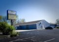 A wide outdoor shot captures a single-story commercial building with light blue vertical metal siding and a dark shingled gabled roof. In the foreground, an asphalt parking lot with faded white parking lines stretches across the frame. On the left, a tall pole sign displays the words "PLYMOUTH COLUMBUS CLUB" in dark text on a white background. Below it, an electronic message board reads "Welcome Lakes Magazine!" in blue and green text against a yellow digital background. A single dark sedan is parked near the side entrance of the building, and green trees and bushes border the property under a bright, clear sky.