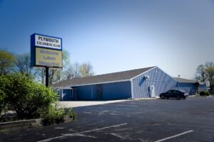A wide outdoor shot captures a single-story commercial building with light blue vertical metal siding and a dark shingled gabled roof. In the foreground, an asphalt parking lot with faded white parking lines stretches across the frame. On the left, a tall pole sign displays the words "PLYMOUTH COLUMBUS CLUB" in dark text on a white background. Below it, an electronic message board reads "Welcome Lakes Magazine!" in blue and green text against a yellow digital background. A single dark sedan is parked near the side entrance of the building, and green trees and bushes border the property under a bright, clear sky.