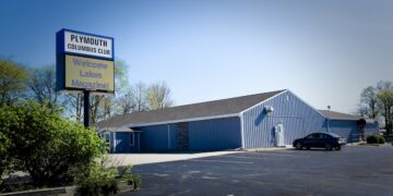 A wide outdoor shot captures a single-story commercial building with light blue vertical metal siding and a dark shingled gabled roof. In the foreground, an asphalt parking lot with faded white parking lines stretches across the frame. On the left, a tall pole sign displays the words "PLYMOUTH COLUMBUS CLUB" in dark text on a white background. Below it, an electronic message board reads "Welcome Lakes Magazine!" in blue and green text against a yellow digital background. A single dark sedan is parked near the side entrance of the building, and green trees and bushes border the property under a bright, clear sky.