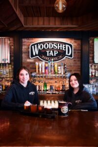 An eye-level shot behind a polished wooden bar counter where two smiling female bartenders stand side-by-side wearing matching dark hoodies. In front of them sits a beer flight sampler paddle with four small glasses and a large, dark ceramic beer stein. Behind them, an exposed brick wall features a row of beer tap handles beneath a large, oval sign that reads "WOODSHED TAP BASS LAKE, IN." Rows of liquor bottles are organized on shelves to the left and right under warm, dim overhead lighting.
