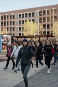 A woman in a grey fleece jacket and beanie waves at the camera while walking in an outdoor plaza. Behind her, a group of people follow along a paved path. A large, multi-story brick building is visible in the background under bright, clear daylight.
