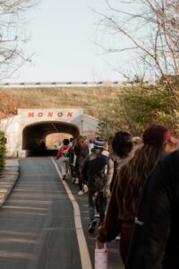 A large group of people is walking along a paved trail toward a concrete tunnel. Above the tunnel's entrance, the word "MONON" is printed in bold red letters. The trail is lined with bare trees, and a road with a guardrail runs along the top of the embankment above the tunnel.