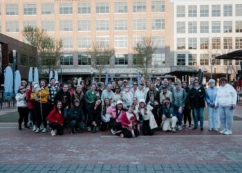 A large, diverse group of about 40 women and two small dogs pose for a group photo in an outdoor urban plaza. They are dressed in casual weekend wear, including hoodies, jackets, and athletic gear. The background features modern brick buildings, a restaurant called "FORK + ALE HOUSE," and the "MIDTOWN" sign in Carmel, Indiana.