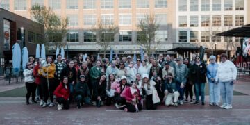 A large, diverse group of about 40 women and two small dogs pose for a group photo in an outdoor urban plaza. They are dressed in casual weekend wear, including hoodies, jackets, and athletic gear. The background features modern brick buildings, a restaurant called "FORK + ALE HOUSE," and the "MIDTOWN" sign in Carmel, Indiana.