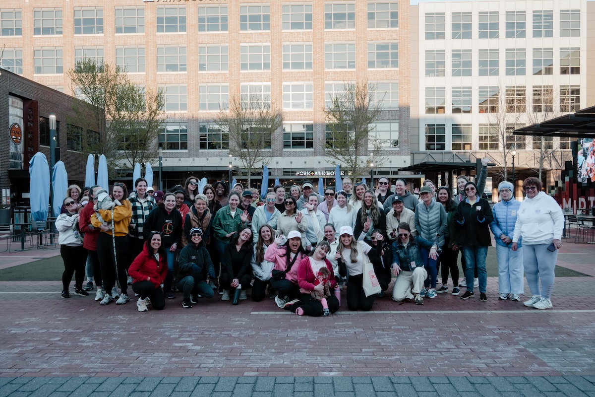 A large, diverse group of about 40 women and two small dogs pose for a group photo in an outdoor urban plaza. They are dressed in casual weekend wear, including hoodies, jackets, and athletic gear. The background features modern brick buildings, a restaurant called "FORK + ALE HOUSE," and the "MIDTOWN" sign in Carmel, Indiana.
