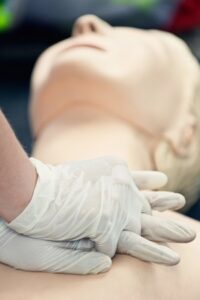 A close-up vertical shot showing a CPR demonstration on a medical mannequin. Two hands wearing white latex gloves are interlocked and positioned in the center of the mannequin's chest to perform compressions. The mannequin's face is visible in the background, tilted slightly upward in a blurred focus.