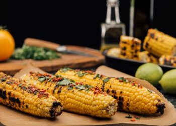 Three ears of grilled corn on the cob rest on brown parchment paper. They are charred in spots and topped with fresh cilantro and red chili flakes. In the blurred background, there are limes, a yellow tomato, chopped herbs on a wooden board, and more corn in a black dish.