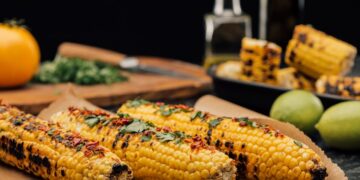 Three ears of grilled corn on the cob rest on brown parchment paper. They are charred in spots and topped with fresh cilantro and red chili flakes. In the blurred background, there are limes, a yellow tomato, chopped herbs on a wooden board, and more corn in a black dish.