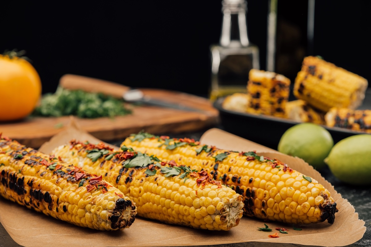 Three ears of grilled corn on the cob rest on brown parchment paper. They are charred in spots and topped with fresh cilantro and red chili flakes. In the blurred background, there are limes, a yellow tomato, chopped herbs on a wooden board, and more corn in a black dish.