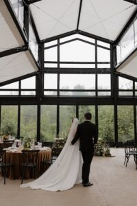 A bride and groom are seen from behind, walking through a modern, glass-walled reception hall. The bride wears a long white gown with a flowing train and a floor-length veil. The groom is in a classic black tuxedo. They are walking toward a large wall of windows overlooking a green forest. In the foreground, a round dining table with a copper-colored cloth and floral centerpieces is visible.