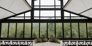 An indoor wedding ceremony space with a modern, industrial feel. Large floor-to-ceiling glass windows with black frames look out onto a lush green forest. Two low, lush floral arrangements sit on the light concrete floor at the altar. Rows of black cross-back chairs face the center. The ceiling features a white fabric canopy with a geometric black metal frame.