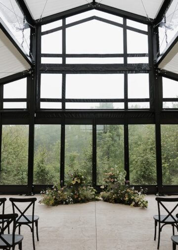 An indoor wedding ceremony space with a modern, industrial feel. Large floor-to-ceiling glass windows with black frames look out onto a lush green forest. Two low, lush floral arrangements sit on the light concrete floor at the altar. Rows of black cross-back chairs face the center. The ceiling features a white fabric canopy with a geometric black metal frame.