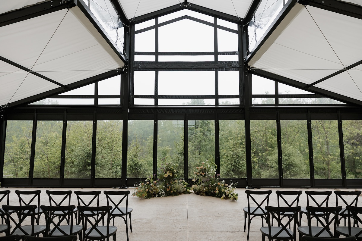 An indoor wedding ceremony space with a modern, industrial feel. Large floor-to-ceiling glass windows with black frames look out onto a lush green forest. Two low, lush floral arrangements sit on the light concrete floor at the altar. Rows of black cross-back chairs face the center. The ceiling features a white fabric canopy with a geometric black metal frame.