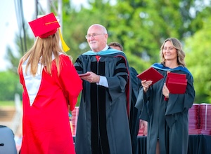 A graduate in a red cap and gown stands on stage, facing away from the camera. She is receiving a diploma from a man in a black academic robe with red and blue doctoral hooding. A woman in similar academic regalia stands next to him, holding a stack of red diploma covers. The outdoor ceremony takes place during the day with green trees in the background.