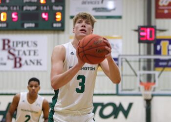 A close-up shot of a male college basketball player wearing a white jersey with the number 3 and "HUNTINGTON" printed across the front. He is focusing intently on the basket, holding a leather basketball in both hands directly in front of his face as he prepares to take a free throw. In the blurred background, another teammate wearing number 2 stands on the court, a scoreboard displays game stats, and a basketball hoop is visible under the gym lighting.