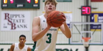A close-up shot of a male college basketball player wearing a white jersey with the number 3 and "HUNTINGTON" printed across the front. He is focusing intently on the basket, holding a leather basketball in both hands directly in front of his face as he prepares to take a free throw. In the blurred background, another teammate wearing number 2 stands on the court, a scoreboard displays game stats, and a basketball hoop is visible under the gym lighting.