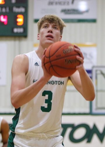 A close-up shot of a male college basketball player wearing a white jersey with the number 3 and "HUNTINGTON" printed across the front. He is focusing intently on the basket, holding a leather basketball in both hands directly in front of his face as he prepares to take a free throw. In the blurred background, another teammate wearing number 2 stands on the court, a scoreboard displays game stats, and a basketball hoop is visible under the gym lighting.