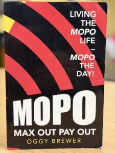 A close-up shot shows the front cover of a book titled "MOPO: Max Out Pay Out" by Oggy Brewer. The cover features a bold, graphic design with a black background and curved red stripes radiating from the bottom left corner. In the upper right, white text reads "Living the MOPO Life – MOPO the Day!" The word "MOPO" is printed in large, thick white letters in the center, with "MAX OUT PAY OUT" and the author's name in smaller white font below it.