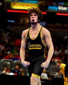 A high school wrestler in a black and gold "Cowan" singlet flexes both arms and shouts in celebration on a wrestling mat. He wears black headgear, and a blurred crowd sits in the arena background beneath brightly lit advertisements.