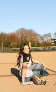 A smiling young woman with long wavy brown hair sits on a dirt softball infield. She wears a white jersey with black sleeves and blue jeans, leaning back slightly while looking at the camera. A softball bat and a leather glove holding a softball rest on the dirt next to her under a clear blue sky.