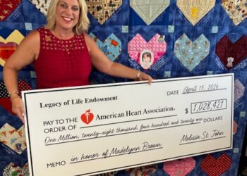 A woman in a red dress smiles brightly while holding a large ceremonial check for $1,028,421 made out to the American Heart Association. The check is dated April 15, 2026, and features the memo "in honor of Madelynn Brown." Behind her is a large blue quilt adorned with numerous colorful, patterned heart patches, some of which contain text and small photos.