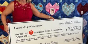 A woman in a red dress smiles brightly while holding a large ceremonial check for $1,028,421 made out to the American Heart Association. The check is dated April 15, 2026, and features the memo "in honor of Madelynn Brown." Behind her is a large blue quilt adorned with numerous colorful, patterned heart patches, some of which contain text and small photos.