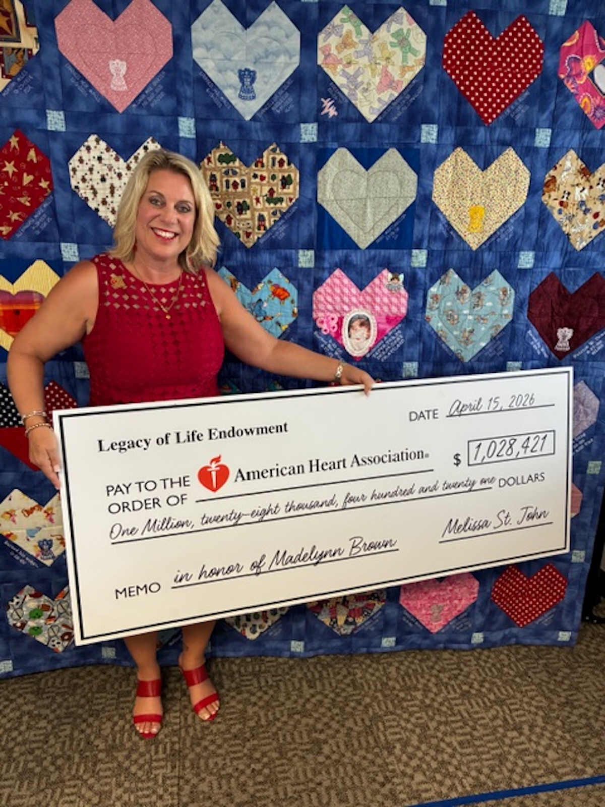 A woman in a red dress smiles brightly while holding a large ceremonial check for $1,028,421 made out to the American Heart Association. The check is dated April 15, 2026, and features the memo "in honor of Madelynn Brown." Behind her is a large blue quilt adorned with numerous colorful, patterned heart patches, some of which contain text and small photos.
