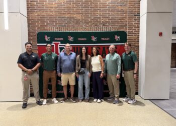 Seven people stand smiling in a row against a brick wall and a green banner that reads "WILDCATS." The banner features a red and white "LN" logo. The group is dressed in business casual and school spirit attire, including green polos and sweaters.