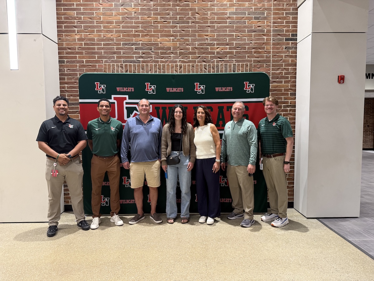 Seven people stand smiling in a row against a brick wall and a green banner that reads "WILDCATS." The banner features a red and white "LN" logo. The group is dressed in business casual and school spirit attire, including green polos and sweaters.