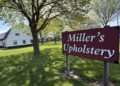 A bright daytime shot of a large burgundy sign on wooden posts that reads "Miller's Upholstery" in white cursive lettering. The sign sits in the foreground on a lush green lawn shaded by a mature tree with fresh spring leaves. In the left background, a large two-story white barn with a dark roof and small square windows is visible. The manicured grass property stretches back toward a line of flowering trees under a clear, bright sky.