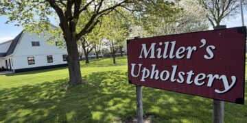 A bright daytime shot of a large burgundy sign on wooden posts that reads "Miller's Upholstery" in white cursive lettering. The sign sits in the foreground on a lush green lawn shaded by a mature tree with fresh spring leaves. In the left background, a large two-story white barn with a dark roof and small square windows is visible. The manicured grass property stretches back toward a line of flowering trees under a clear, bright sky.