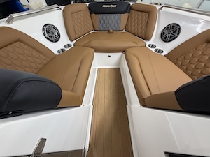 A close-up view looking into the bow seating area of a modern white boat, featuring premium tan upholstery with diamond-patterned stitching and dark gray accents. The u-shaped lounge seating wraps around a central faux-teak walkways floor. Built into the white fiberglass hull sides are circular marine stereo speakers with silver grilles, alongside several stainless steel cup holders.