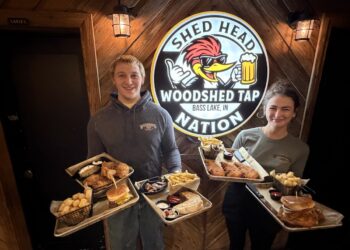 A man and a woman stand smiling indoors against a rustic wooden wall, each holding two metal trays piled with pub food. The man wears a gray hoodie and the woman wears a gray t-shirt, both featuring a "Woodshed Tap" logo. Between them is a large circular sign that reads "SHED HEAD NATION" and "WOODSHED TAP BASS LAKE, IN," featuring a cartoon woodpecker wearing sunglasses and holding a beer mug. The food trays contain burgers, fries, tater tots, chicken tenders, and quesadillas.