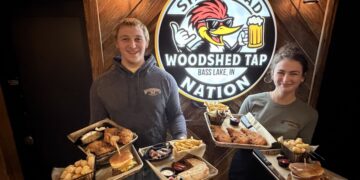 A man and a woman stand smiling indoors against a rustic wooden wall, each holding two metal trays piled with pub food. The man wears a gray hoodie and the woman wears a gray t-shirt, both featuring a "Woodshed Tap" logo. Between them is a large circular sign that reads "SHED HEAD NATION" and "WOODSHED TAP BASS LAKE, IN," featuring a cartoon woodpecker wearing sunglasses and holding a beer mug. The food trays contain burgers, fries, tater tots, chicken tenders, and quesadillas.