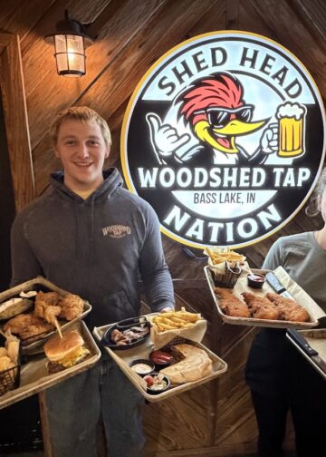 A man and a woman stand smiling indoors against a rustic wooden wall, each holding two metal trays piled with pub food. The man wears a gray hoodie and the woman wears a gray t-shirt, both featuring a "Woodshed Tap" logo. Between them is a large circular sign that reads "SHED HEAD NATION" and "WOODSHED TAP BASS LAKE, IN," featuring a cartoon woodpecker wearing sunglasses and holding a beer mug. The food trays contain burgers, fries, tater tots, chicken tenders, and quesadillas.
