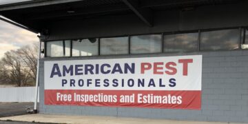 A large vinyl sign hangs on the side of a grey cinder block commercial building with wide glass windows. The banner reads "American Pest Professionals" in dark blue and red text on a white background, with a red bottom section stating "Free Inspections and Estimates." An asphalt driveway and bare winter trees under a cloudy sky are visible in the foreground and background.
