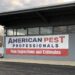 A large vinyl sign hangs on the side of a grey cinder block commercial building with wide glass windows. The banner reads "American Pest Professionals" in dark blue and red text on a white background, with a red bottom section stating "Free Inspections and Estimates." An asphalt driveway and bare winter trees under a cloudy sky are visible in the foreground and background.