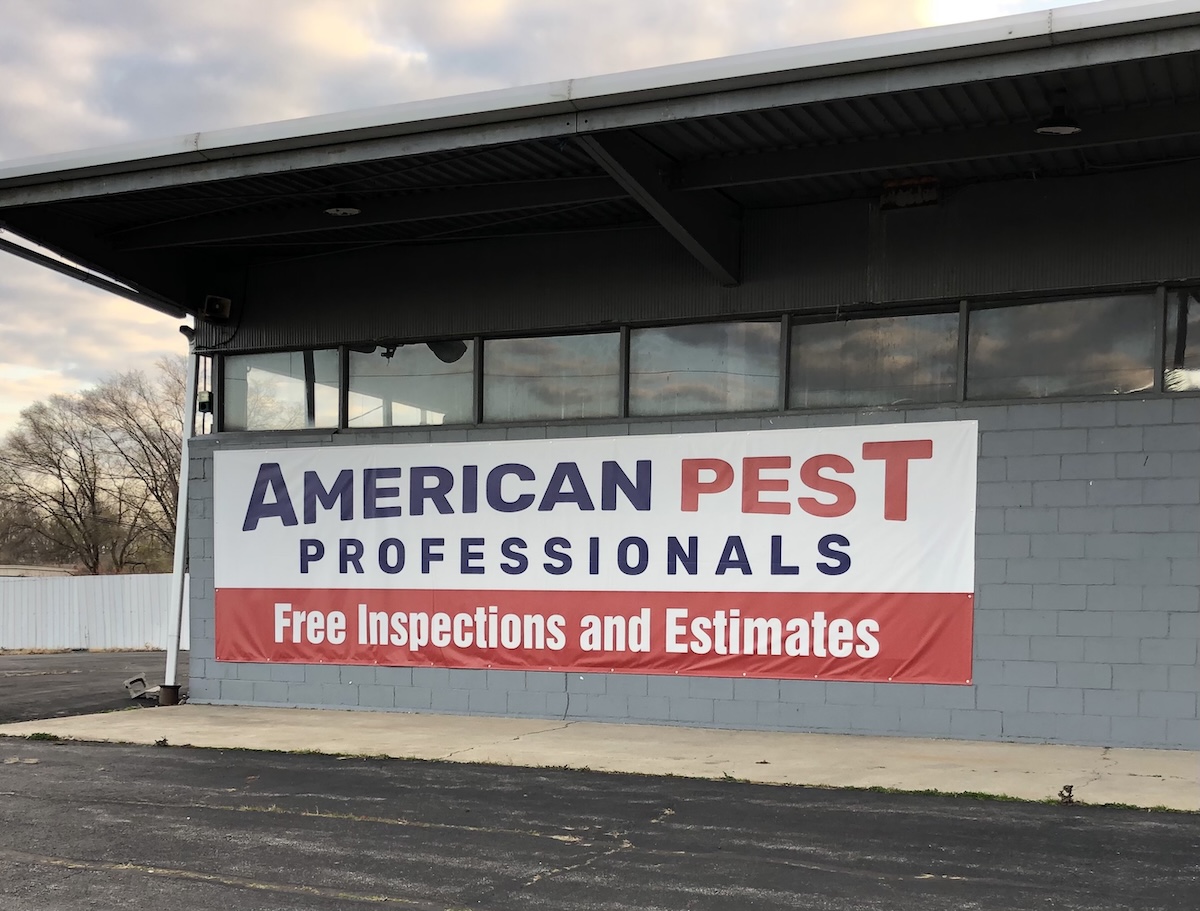 A large vinyl sign hangs on the side of a grey cinder block commercial building with wide glass windows. The banner reads "American Pest Professionals" in dark blue and red text on a white background, with a red bottom section stating "Free Inspections and Estimates." An asphalt driveway and bare winter trees under a cloudy sky are visible in the foreground and background.