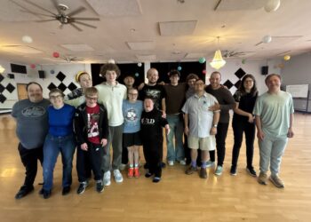 A large, diverse group of about 15 young adults stands together on a wooden dance floor in a studio. They are all smiling and have their arms around each other for a group photo. The studio is decorated with colorful paper lanterns, and black-and-white checkered diamond patterns are on the back wall. The individuals are dressed casually in t-shirts, hoodies, and jeans.