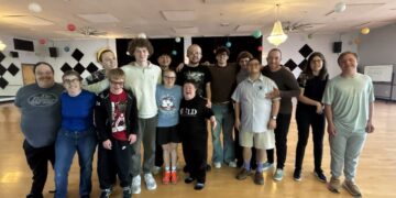 A large, diverse group of about 15 young adults stands together on a wooden dance floor in a studio. They are all smiling and have their arms around each other for a group photo. The studio is decorated with colorful paper lanterns, and black-and-white checkered diamond patterns are on the back wall. The individuals are dressed casually in t-shirts, hoodies, and jeans.