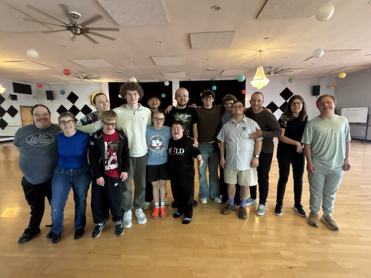 A large, diverse group of about 15 young adults stands together on a wooden dance floor in a studio. They are all smiling and have their arms around each other for a group photo. The studio is decorated with colorful paper lanterns, and black-and-white checkered diamond patterns are on the back wall. The individuals are dressed casually in t-shirts, hoodies, and jeans.