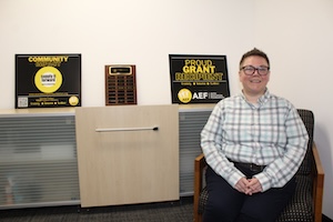 A woman in a plaid shirt and glasses sits in an office chair, smiling at the camera. Behind her, on a counter, are two black signs with yellow text and a wooden plaque. One sign says "COMMUNITY PARTNER" and the other says "PROUD GRANT RECIPIENT" with the Anderson Education Foundation (AEF) logo. The room has neutral white walls and modern office furniture.