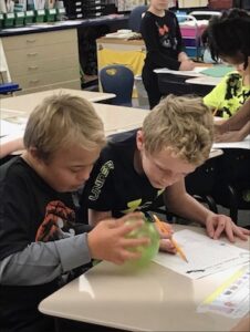 Two young boys sit at a classroom desk, focused on a science experiment. One boy holds a translucent green sphere while the other writes on a worksheet with a pencil. In the background, other students are visible at their desks in a well-organized classroom with cabinets and storage bins.