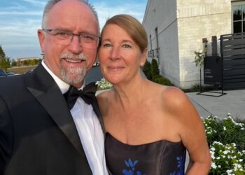 A smiling couple poses for a selfie outdoors at a formal event. The man on the left wears a black tuxedo, a white shirt, a black bow tie, and glasses. The woman on the right wears a strapless black dress with blue floral patterns. In the background, a white brick building, green landscaping, and a black sign with the names "Emma & Patrick" are visible under a clear blue sky.