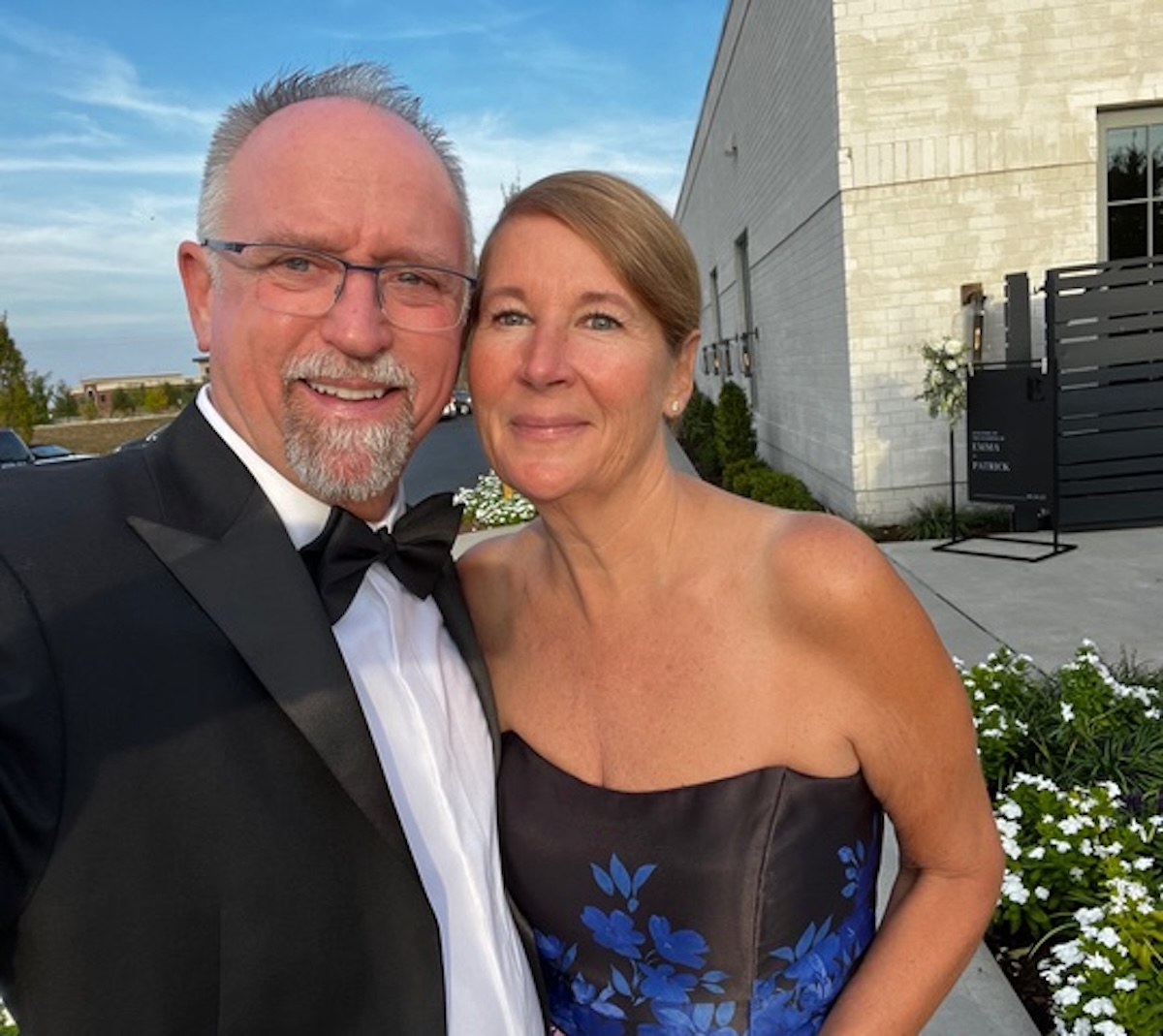 A smiling couple poses for a selfie outdoors at a formal event. The man on the left wears a black tuxedo, a white shirt, a black bow tie, and glasses. The woman on the right wears a strapless black dress with blue floral patterns. In the background, a white brick building, green landscaping, and a black sign with the names "Emma & Patrick" are visible under a clear blue sky.