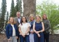 A group of six professionally dressed adults—five women and one man—pose together outdoors in front of a thick, textured tree trunk. They stand close together smiling, with tall evergreen trees and leafy green bushes creating a natural background.